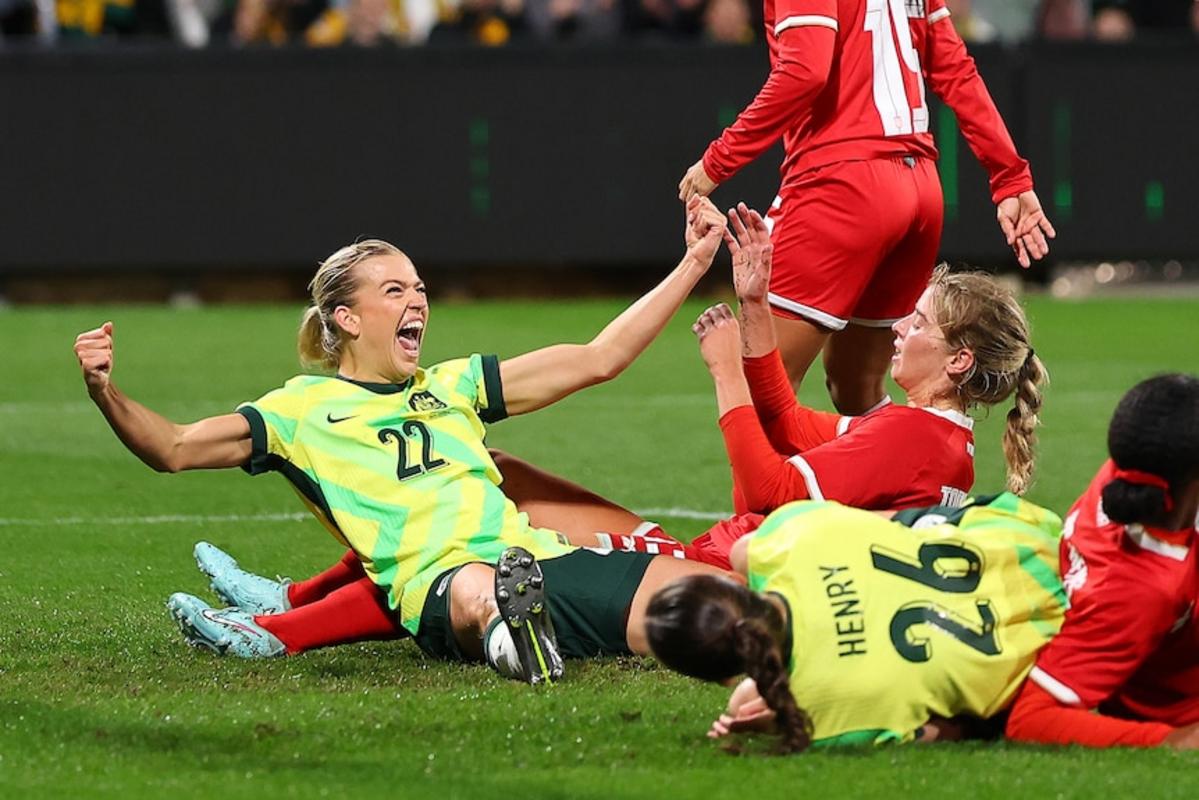 Một cầu thủ bóng đá mặc áo vàng và xanh ngồi xuống sân và giơ tay chiến thắng